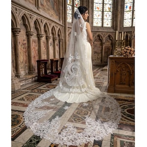 Pode incluir: Uma noiva com um vestido de noiva branco e véu está em uma igreja. O véu é longo e apresenta detalhes intrincados de renda e uma imagem religiosa. A igreja tem vitrais e detalhes arquitetônicos ornamentados.