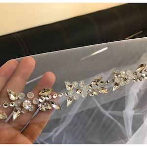 May include: Close-up of a white tulle fabric with a silver beaded trim. The trim features a repeating pattern of small, clear crystals and silver beads arranged in a floral design.