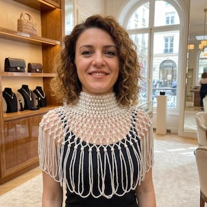May include: A high-neck pearl capelet featuring a structured collar of multiple rows of white pearls, transitioning into a delicate lattice pattern of interconnected pearl strands that drape over the shoulders, ending in a fringe of hanging pearl loops.