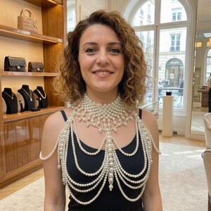 May include: A multi-layered pearl body necklace featuring a high-neck collar design with intricate draping strands of white pearls that extend over the shoulders and across the chest, creating a cascading effect over a simple black tank top.