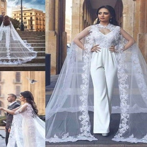 Op de afbeelding: Een witte mouwloze jumpsuit met een kanten lijfje, een hoge halslijn en een uitgesneden detail. Het kledingstuk bevat een lange, doorschijnende cape tot op de grond, versierd met ingewikkelde witte bloemenkant langs de randen en zoom.