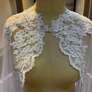 Op de afbeelding: Een witte kanten bruidscape met bloemenborduurwerk. Het delicate kant heeft gedetailleerde bloemen- en bladerpatronen. De cape is over een mannequin gedrapeerd en toont het ingewikkelde ontwerp en de transparante stof.