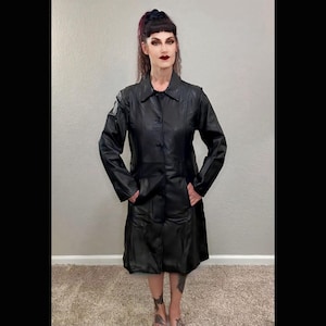 Puede incluir: Un abrigo de cuero negro con botones, mangas largas y cuello. El abrigo tiene bolsillos y llega hasta la pantorrilla. El abrigo lo lleva una persona.