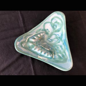 Puede incluir: Un cenicero triangular con acabado brillante. El cenicero presenta una mezcla de colores turquesa, azul y blanco, con un patrón arremolinado. Tiene un reposo para cigarrillos incorporado. El cenicero es un artículo vintage.