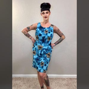 Può includere: Abito senza maniche, lunghezza al ginocchio, con motivo floreale blu. L'abito presenta uno scollo tondo e una silhouette aderente. Il design floreale include fiori blu, beige e bianchi su sfondo azzurro. L'abito è realizzato in tessuto leggero.