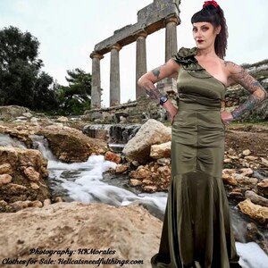 May include: A woman wearing a one-shoulder, olive green satin dress with a large flower detail on the shoulder. She is standing in front of an ancient stone structure with a small waterfall in the foreground. The text "Photography: HKMorales" and "Clothes for Sale: Hellcatsneedfulthings.com" is at the bottom of the image.