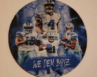 May include: A round, blue and white fabric coaster featuring a photo of four football players in blue and white uniforms. The text "WE DEM BOYZ" is printed on the bottom of the coaster.