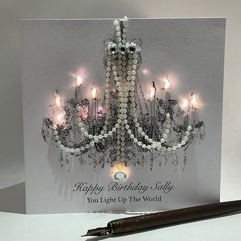 May include: A birthday card with a black and white chandelier design, adorned with pearls and crystals. The card's message is "Happy Birthday Sally, You Light Up The World". A vintage pen is in the foreground.