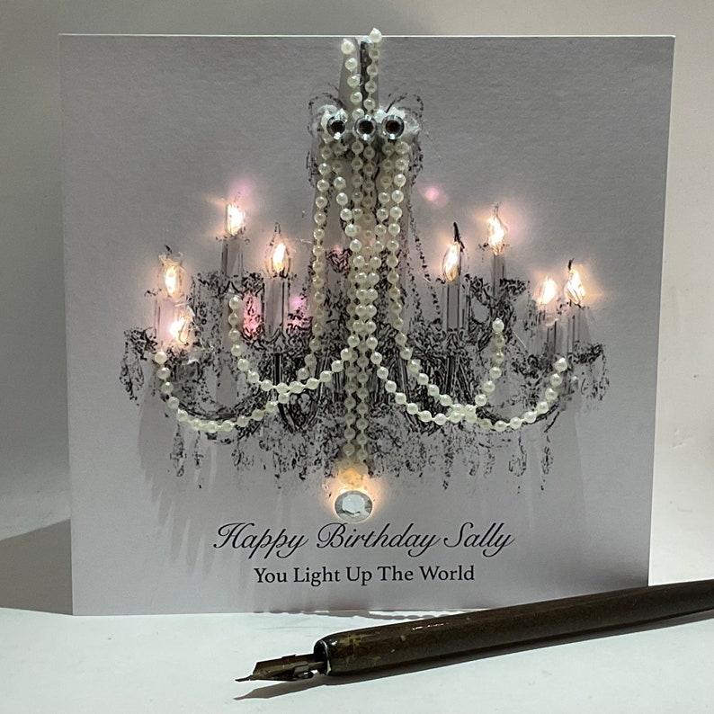 May include: A white birthday card with a black and white chandelier design, embellished with pearls and crystals. The card reads "Happy Birthday Sally, You Light Up The World". A pen is in the foreground.