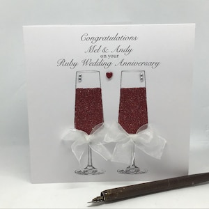Pode incluir: Um cartão branco com um coração vermelho e duas taças de champanhe cheias de glitter vermelho. O cartão diz "Parabéns Mel & Andy pelo seu aniversário de casamento de rubi".