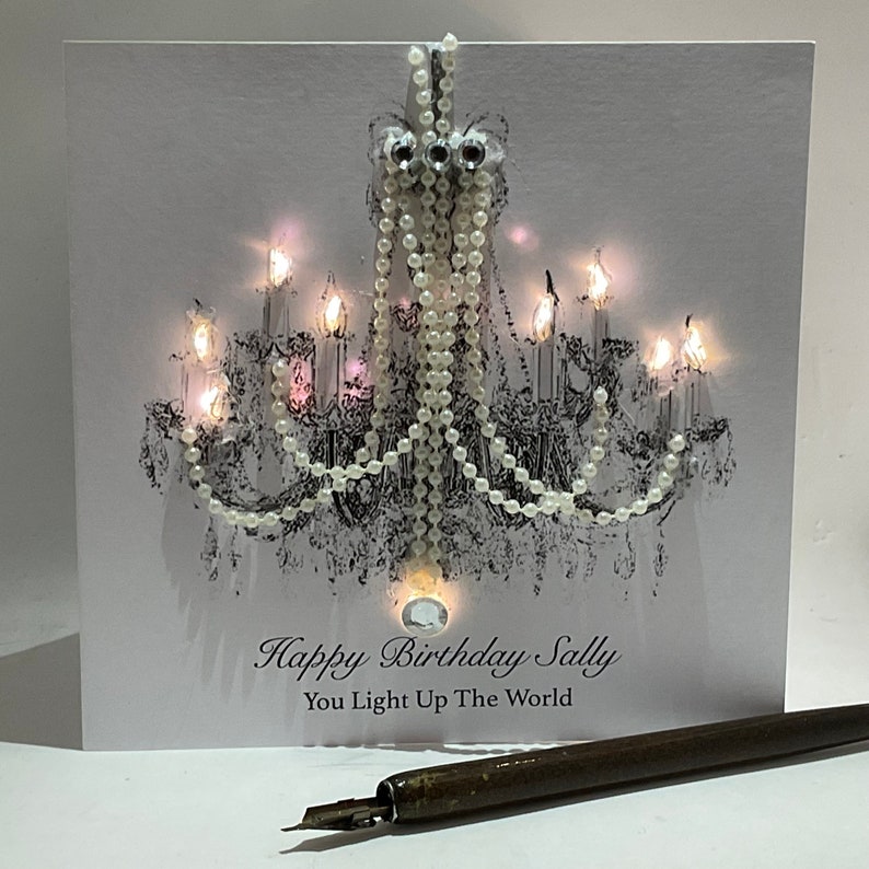 May include: A white birthday card with a chandelier design, adorned with pearls and rhinestones. The card reads "Happy Birthday Sally, You Light Up The World." A vintage pen is positioned in the foreground.