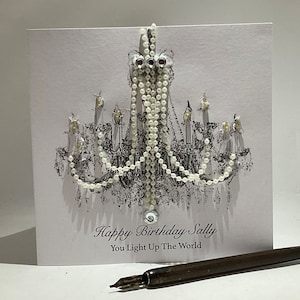 May include: A white birthday card featuring a black and white chandelier design, adorned with white beads and crystals. The card has the text "Happy Birthday Sally, You Light Up The World". A pen is in the foreground.