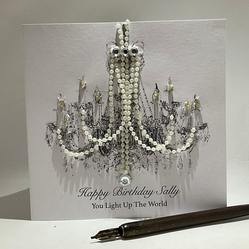 May include: A white birthday card featuring a black and white chandelier design, adorned with white beads and crystals. The card has the text "Happy Birthday Sally, You Light Up The World". A pen is in the foreground.
