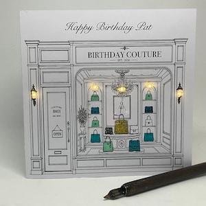 May include: A square greeting card featuring a black and white line drawing of a boutique storefront labelled BIRTHDAY COUTURE EST. 2026. The shop window displays various handbags on shelves, illuminated by small integrated warm yellow LED lights.