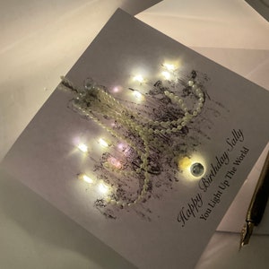 May include: A birthday card with the message "Happy Birthday Sally, You Light Up The World." The card features a chandelier design with small lights and a string of pearls. A pen is visible in the bottom right corner.