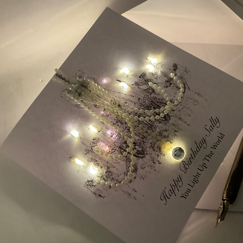 May include: A birthday card with the message "Happy Birthday Sally, You Light Up The World." The card features a chandelier design with small lights and a string of pearls. A pen is visible in the bottom right corner.