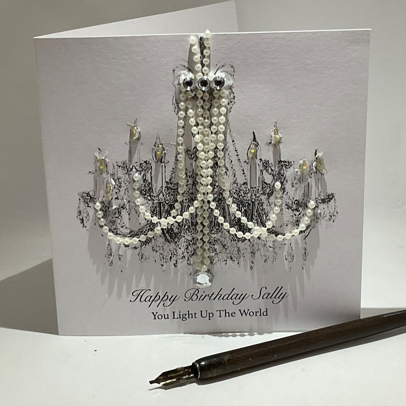 May include: A white birthday card with a black and white chandelier design, adorned with pearl and crystal details. The card says "Happy Birthday Sally, You Light Up The World." A vintage pen is placed on the surface.