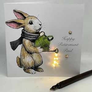 May include: A white card with a brown rabbit holding a green watering can. The rabbit is wearing a black and white scarf. The card has the text "Happy Retirement Paul" on it.