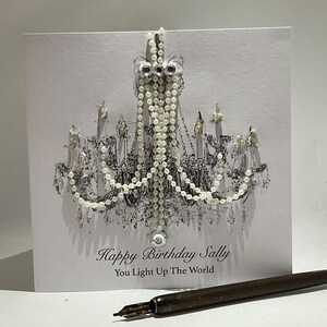 May include: A birthday card with a chandelier design. The card is white with a black and white illustration of a chandelier and the text "Happy Birthday Sally, You Light Up The World". A pen is in the foreground.