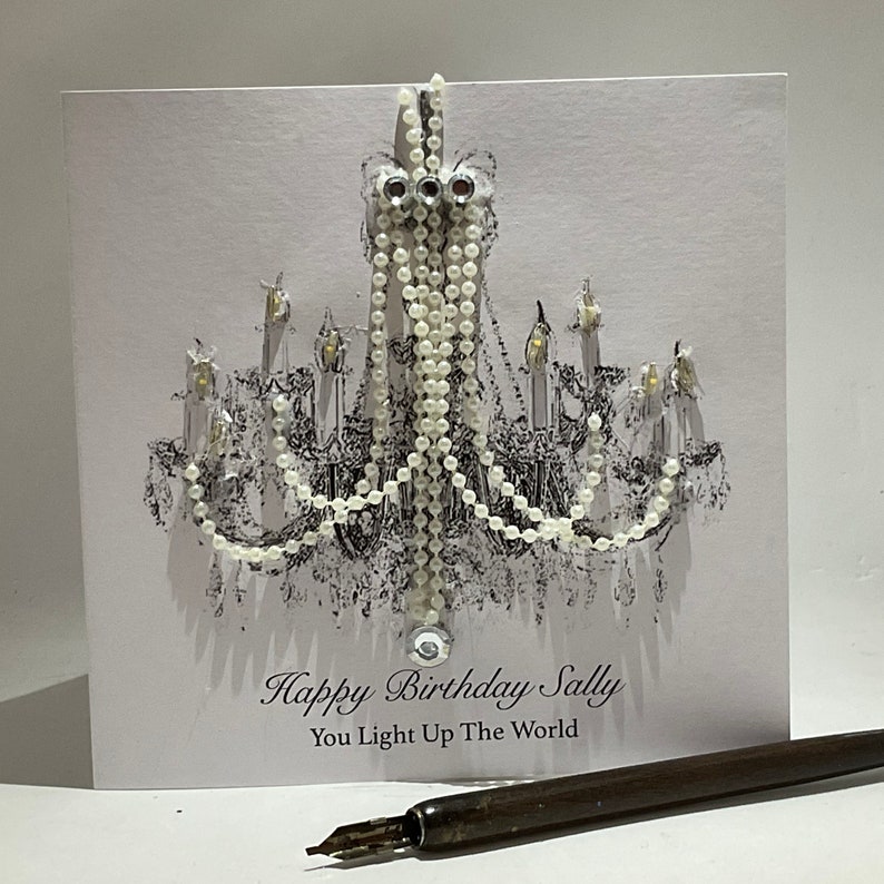 May include: A birthday card with a chandelier design. The card is white with a black and white illustration of a chandelier and the text "Happy Birthday Sally, You Light Up The World". A pen is in the foreground.