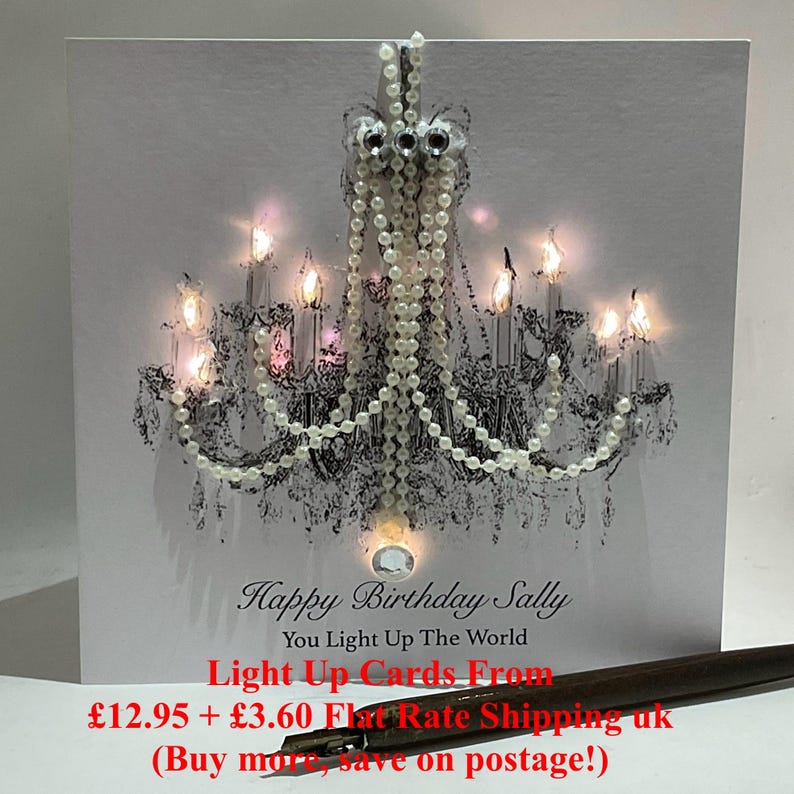 May include: A white birthday card with a black and white chandelier design, adorned with pearl detailing and small lights. The card's text reads "Happy Birthday Sally, You Light Up The World".