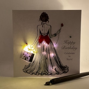 May include: A white card with a black and white illustration of a woman in a gown holding a gift box with lights. The text "Happy Birthday Celebrate in Style!" is printed on the card.