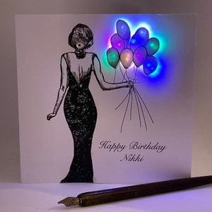 May include: A white card with a black and white illustration of a person in a dress holding a bunch of balloons. The balloons are glowing with multi-coloured lights. The card says "Happy Birthday Nikki".