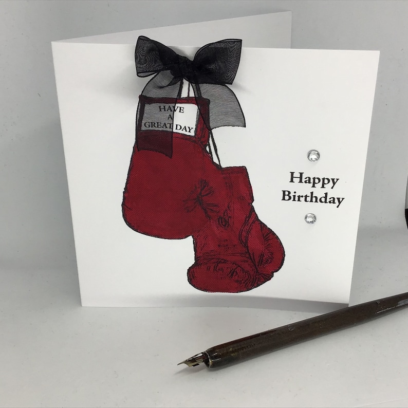 Może przedstawiać: Biała kartka z czerwonym bokserskim rękawem wiszącym na czarnej wstążce. Na kartce widnieje napis "Have a Great Day" i "Happy Birthday".