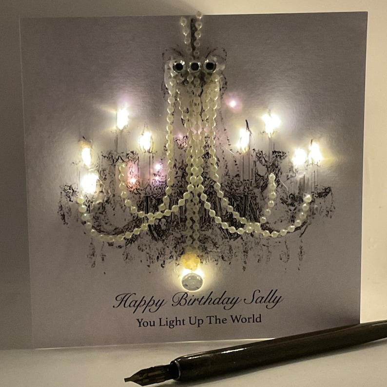 May include: A birthday card with a chandelier design. The card features a black and white illustration of a chandelier with pearl-like beads and small lights. The text "Happy Birthday Sally, You Light Up The World" is printed on the card. A pen rests in the foreground.