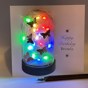 May include: A white card with a glass dome containing a string of colourful lights and black butterfly cutouts. The card reads "Happy Birthday Brenda".