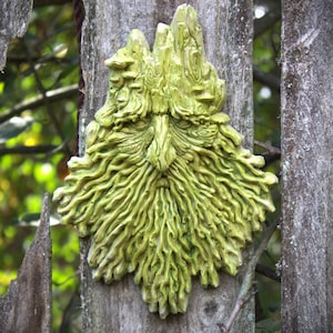 Puede incluir: Una escultura de pared de un rostro con una barba y cabello largos y fluidos, elaborada con una apariencia texturizada similar a la madera en tonos verde claro y amarillo. El diseño presenta patrones intrincados y arremolinados.