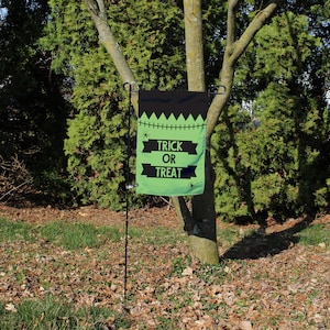 May include: A Halloween garden flag with a black top and green bottom. The flag has a jagged edge design and the words "TRICK OR TREAT" in black. Small spider graphics are also present. The flag is displayed outdoors, attached to a black metal stand.