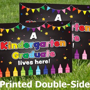 May include: Two black chalkboard signs with colorful decorations. The top sign says "A Kindergarten Graduate!" The bottom sign says "A Kindergarten Graduate lives here!" Both signs have a row of colorful crayons at the bottom.