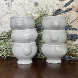 May include: Six white ceramic bowls stacked in two columns of three. Each bowl features a different embossed animal face. The bowls are on a dark wooden surface, with dried lavender and a textured background.