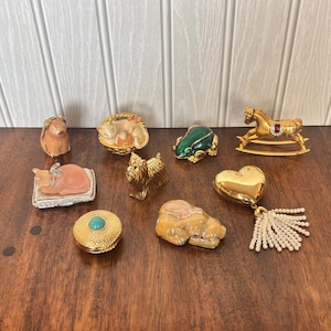 May include: A collection of small decorative figurines including a tan dog, a cat in a basket, a green frog, a gold rocking horse, a cat on a cushion, a gold dog, a round gold box with a turquoise stone, a tan stone animal, and a gold heart with a pearl tassel.