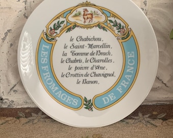 May include: A white ceramic plate with a blue and yellow border. The plate features a goat illustration and text in French that reads: "LES FROMAGES DE FRANCE le Chabichou, le Saint-Marcellin, la Comme de Brach, le Chabris, le Charolles, le poivre d'Ane, le Crottin de Chavignol, le Banon."