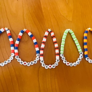 May include: Five colorful beaded bracelets with the words "DETROIT", "PARIS", "MCM", "EUGENE", and "CINCIN" spelled out. The bracelets feature red, white, blue, green, and yellow beads, and are arranged on a wooden surface.
