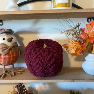 May include: A burgundy crocheted pumpkin sits on a wooden shelf with a small bird figurine and a vase of fall foliage.