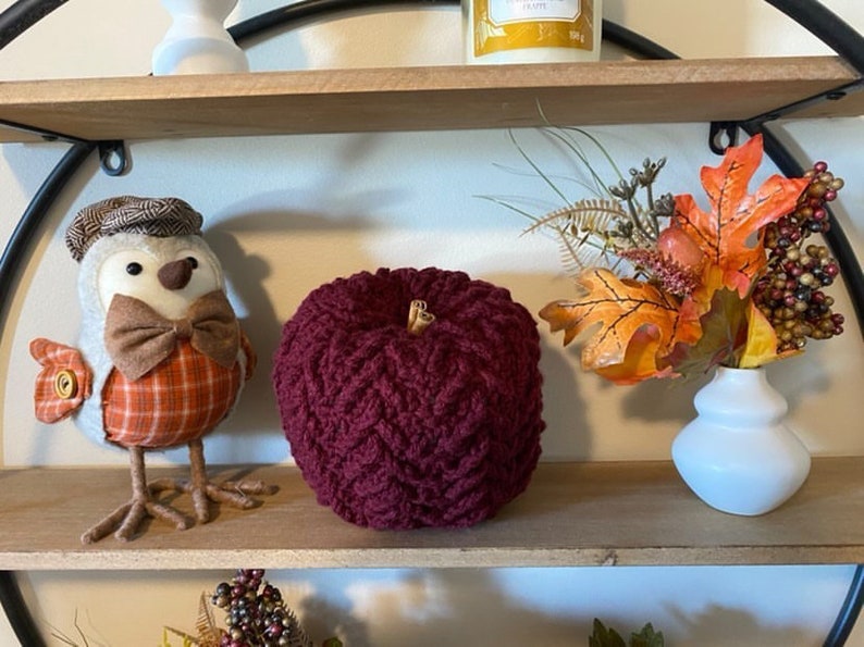 May include: A burgundy crocheted pumpkin sits on a wooden shelf with a small bird figurine and a vase of fall foliage.