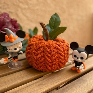 May include: A crocheted orange pumpkin with green leaves sits on a wooden table with two small Halloween-themed figurines.