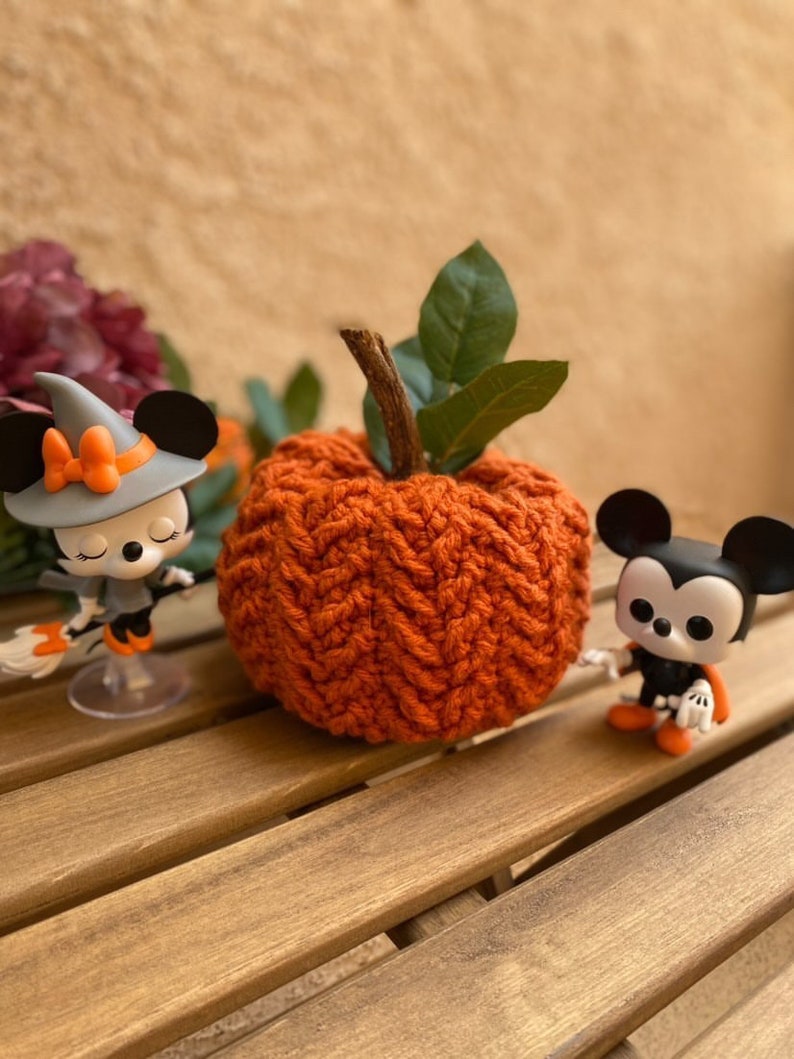 May include: A crocheted orange pumpkin with green leaves sits on a wooden table with two small Halloween-themed figurines.