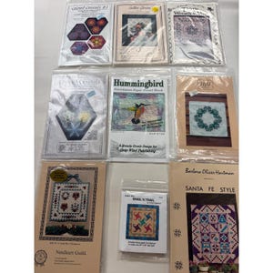 May include: A collection of eight quilting and needlework pattern booklets in clear plastic sleeves. Titles include Colored Crystals, Lattice Garden, Thistle Wreath, Hummingbird, A Little Bit of Baltimore, Snail's Trail, and Santa Fe Style.