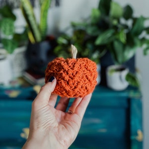 May include: A crocheted orange pumpkin with a textured surface. The pumpkin is being held in a hand.