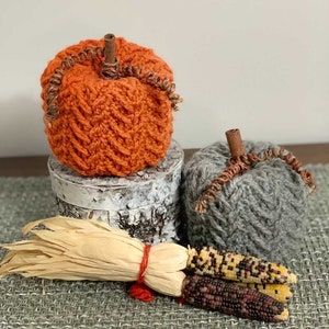 May include: Two knitted pumpkins, one orange and one gray, with a brown stem and a curled twig. The pumpkins are sitting on a wooden surface with a woven mat and a bundle of dried corn husks.