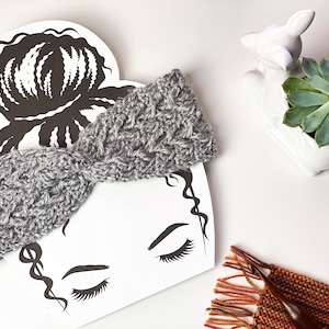 May include: A gray crocheted headband with a twisted design. The headband is displayed on a white background with a black silhouette of a woman's face.
