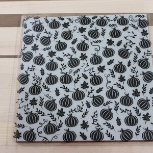 May include: A clear sheet of vinyl with a black and white pattern of pumpkins and leaves. The pumpkins are stylized and have a simple design.