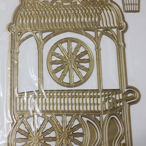 May include: Gold metal die cut of a decorative cart with a roof, two wheels, and a railing. The cart is designed to be used for crafting and scrapbooking.