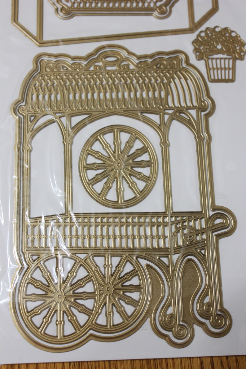 May include: Gold metal die cut of a decorative cart with a roof, two wheels, and a railing. The cart is designed to be used for crafting and scrapbooking.