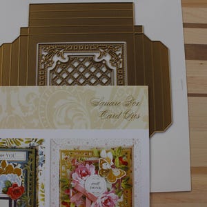 May include: A gold-colored metal die-cutting template for a square box card, featuring an intricate lattice design. The packaging displays the Anna Griffin brand name, the text Square Box Card Dies, and two sample cards decorated with floral motifs.