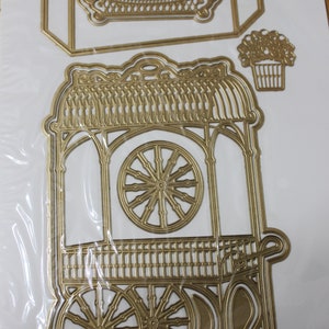 May include: A gold metal die-cut set of a decorative cart with a wheel, a rectangular sign, and a flower pot. The cart has a decorative roof and a lattice-style side.